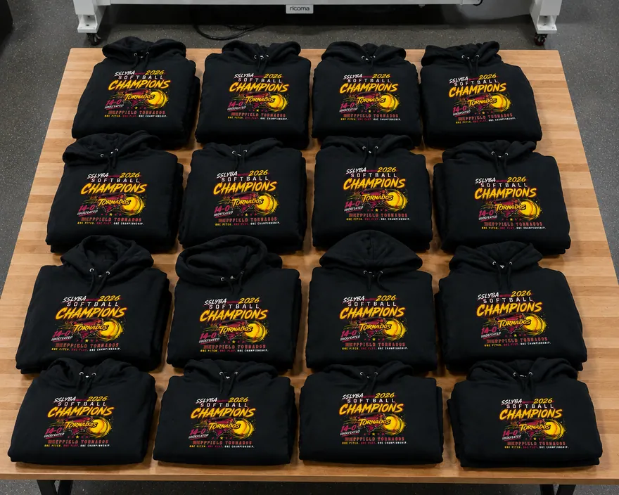 Sixteen black championship hoodies folded and boxed on the packing table, printed for an undefeated softball team
