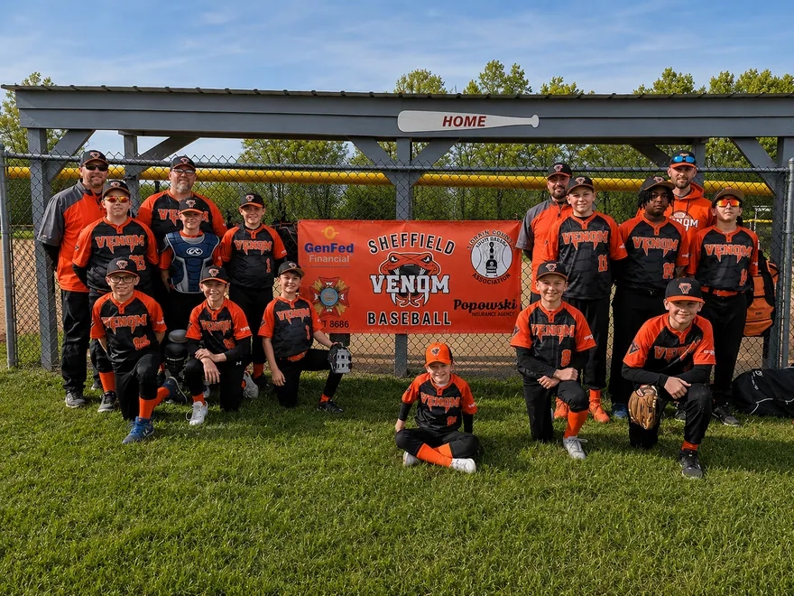 Youth baseball team in black and orange sublimated jerseys, caps, socks and belts in front of their team banner